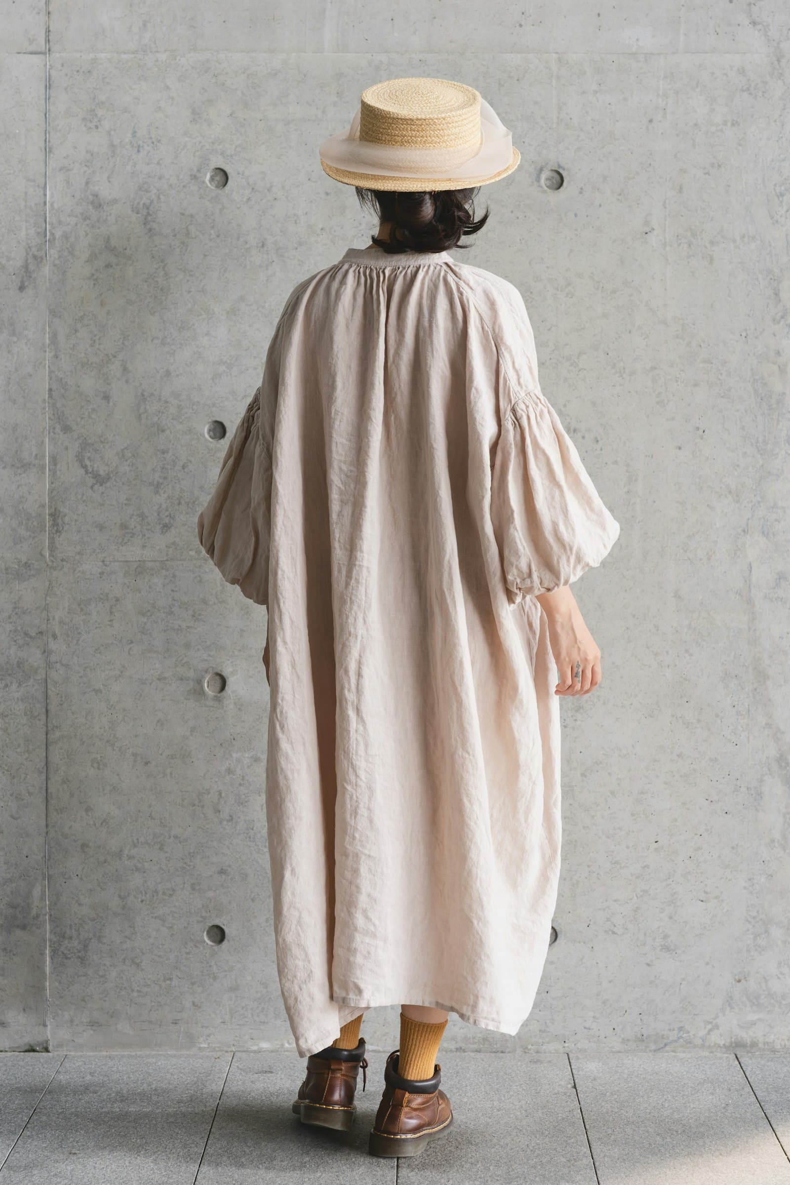 Woman in straw hat and linen dress stands against wall