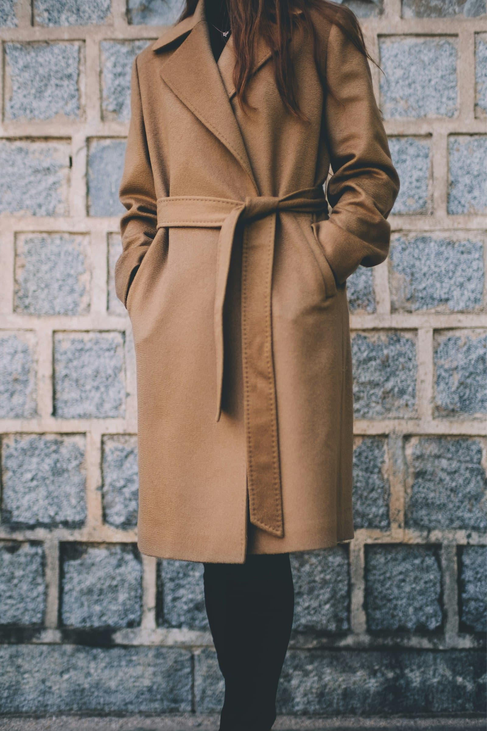 Woman wearing brown single-breasted coat standing in front of gray brick wall